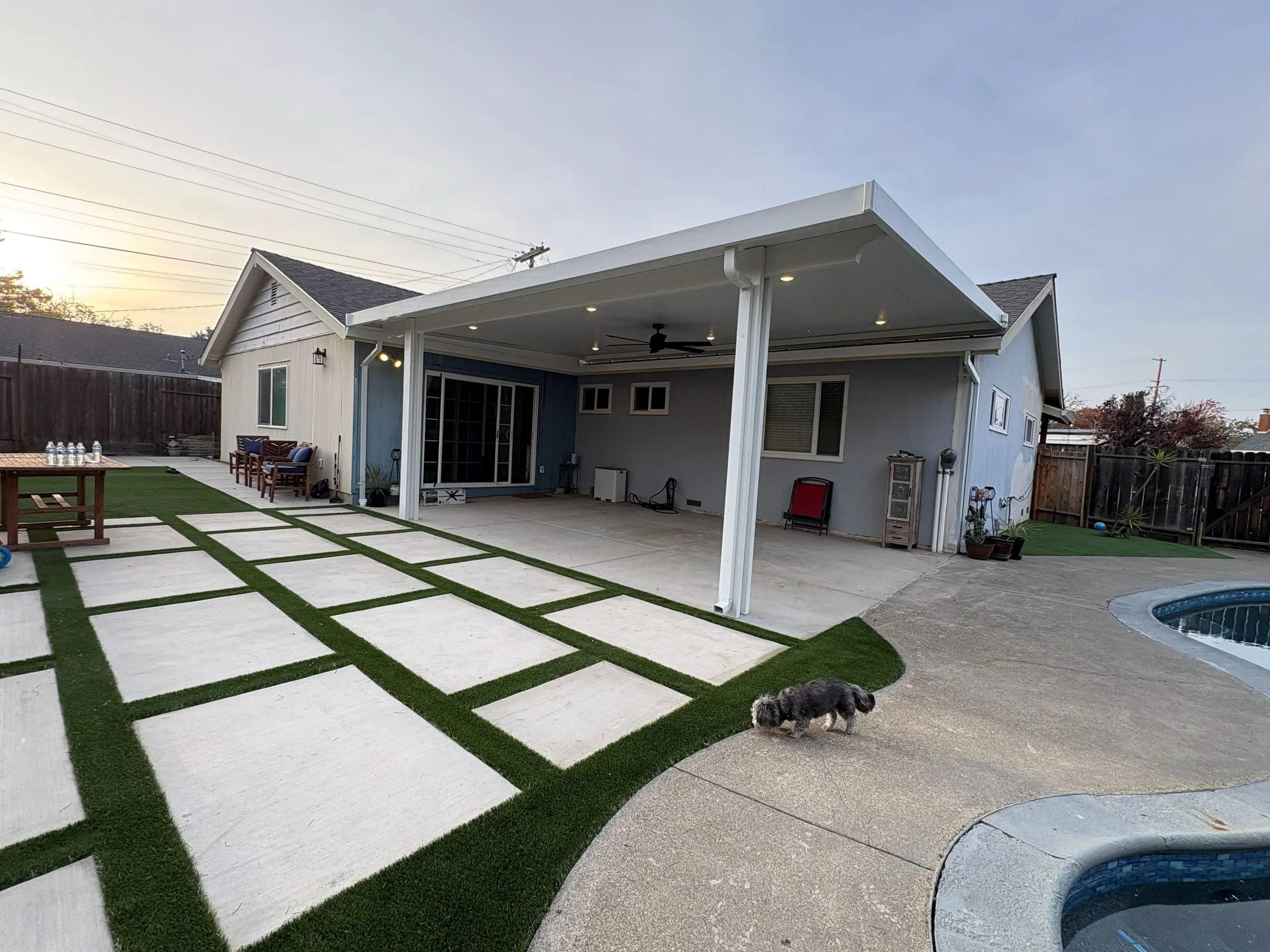 White patio cover with lights beside a pool, concrete pavers and turf strips